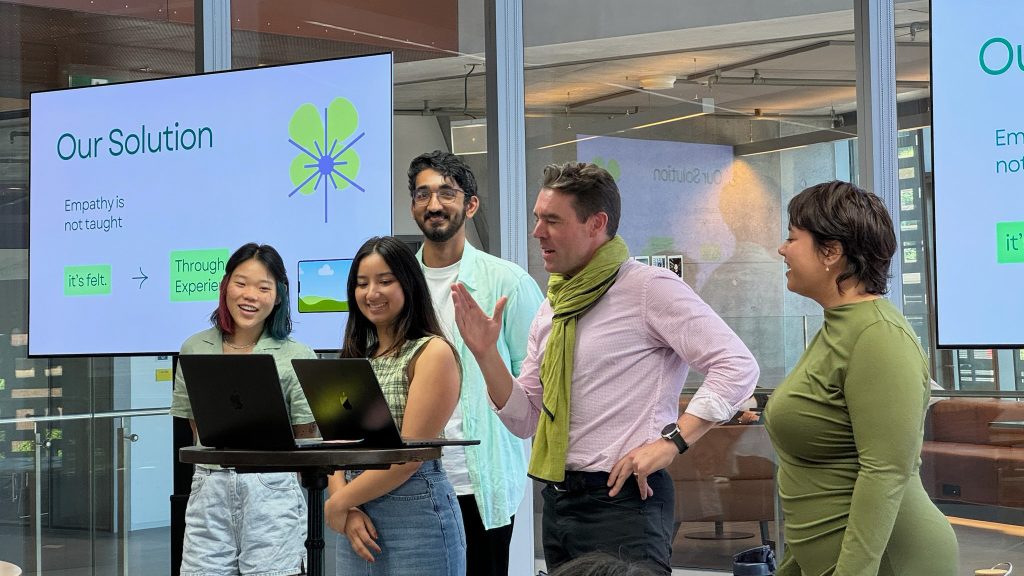 A group of smiling students is standing at a laptop with a presentation screen behind them.