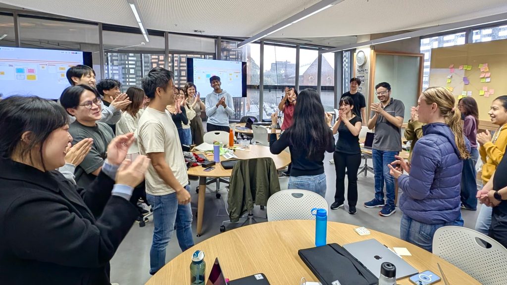 a group of about 15-20 students are standing up in a classroom, smiling and clapping
