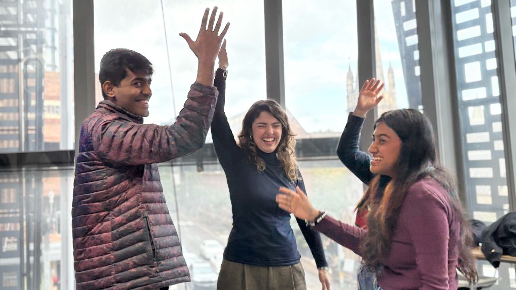 4 students standing with a hand in the air, demonstrating a team handshake exercise