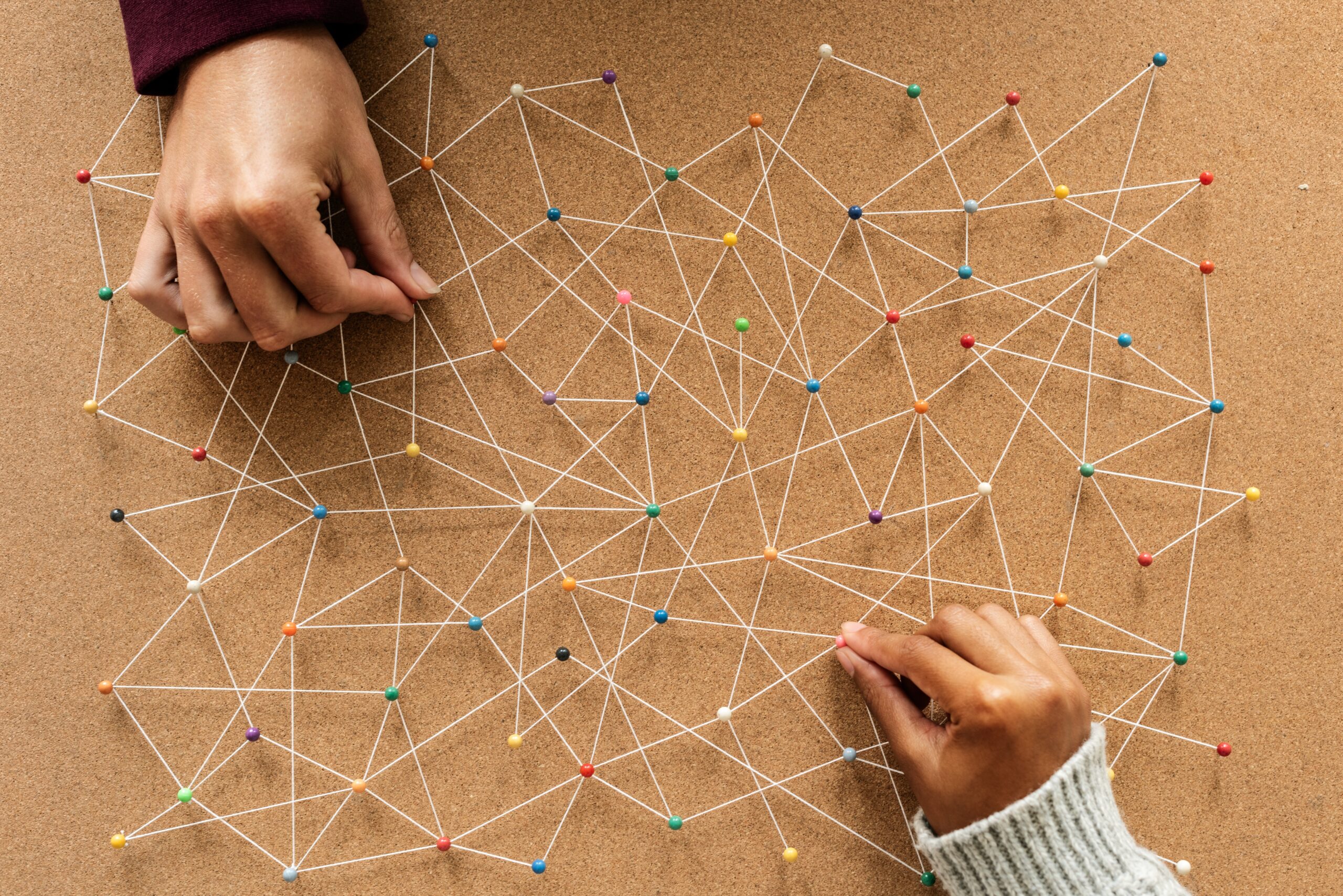 a cork board with a network of pins on it. 2 human hands are adding pins.