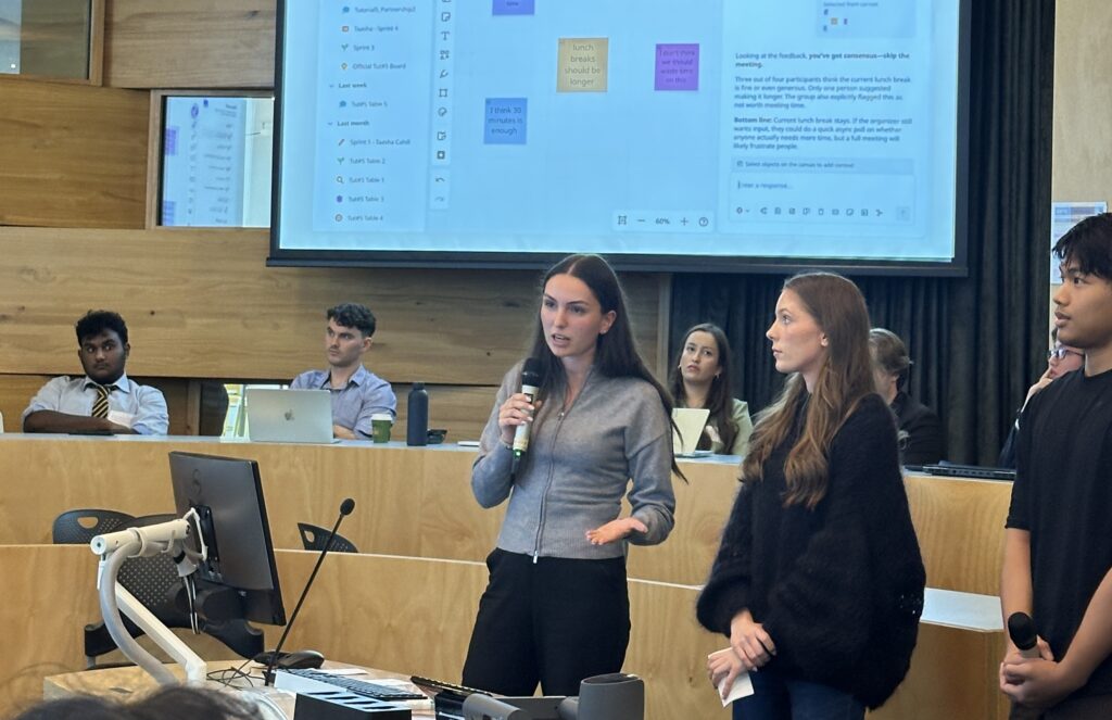 Students pitch their challenge response in front of a Miro Board screen.